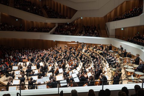 Foto: Großes Universitätsorchester auf einer Bühne in einem Konzertsaal mit Publikum auf mehreren Rängen