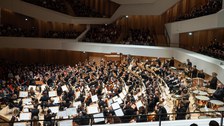 Foto: Großes Universitätsorchester auf einer Bühne in einem Konzertsaal mit Publikum auf mehreren Rängen