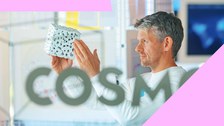 Foto: Eine Person hält ein weißes, würfelförmiges Objekt mit schwarzen Mustern vor eine Glaswand mit der Aufschrift 'COSMO'