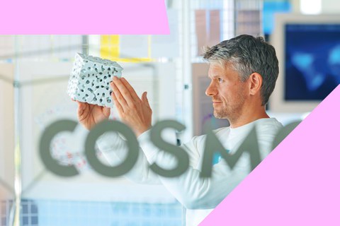 Foto: Eine Person hält ein weißes, würfelförmiges Objekt mit schwarzen Mustern vor eine Glaswand mit der Aufschrift 'COSMO'