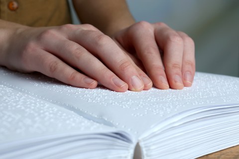 Foto eines Buches mit Brailleschrift, auf dem zwei Hände die Schriftzeichen berühren.