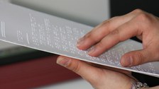 Foto eines Brailleausdrucks. Eine Hand hält das Papier fest, die andere ertastet das Braille.