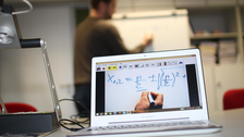 Foto einer beispielhaften Lehrsituation. Im Vordergrund befindet sich rechts ein Laptop, links eine Tafelbildkamera. Im Hintergrund erkennt man schwach eine Person an einer Flipchart, welche darauf zeichnet. Auf dem Bildschirm des Laptops sieht man in Nahaufnahme eine weiße Oberfläche mit einer zeichnenden Hand und einer Formel darüber.