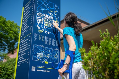 Foto einer Person mit Krücken, die versucht sich mit Hilfe einer Informationskarte auf dem TU Campus zu orientieren