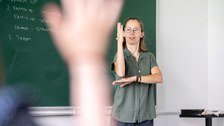 Foto einer Person vor einer Tafel, die etwas in Gebärdensprache erzählt. In der linken Bildhälfte ist eine sich meldende Hand zu sehen. 