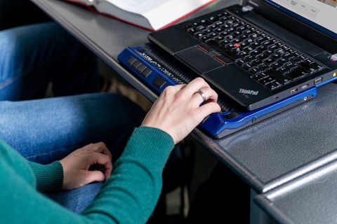 Foto einer Person, deren Arm zu sehen ist. Die Person sitzt an einem Laptop und ihre Hand liegt auf einer Braillezeile.