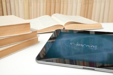 Photo of a table with a tablet in the foreground and four unrecognizable books in the background. The screen of the tablet shows a blackboard surface and the chalk writing »e-learning«.
