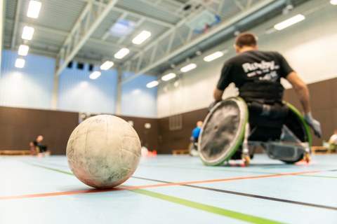Foto einer Sporthalle. Im Vordergrund ein Handball, im Hintergrund unscharf mehrere Rollstuhlfahrer.