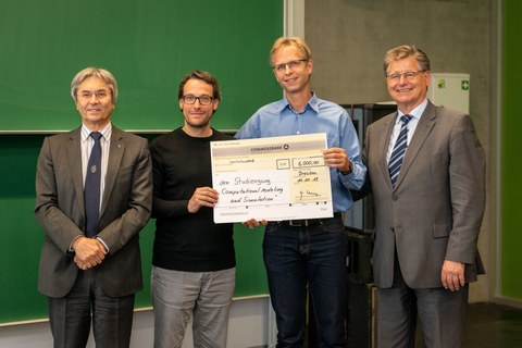 The image shows four men standing in front of a chalkboard. The two in the middle are holding up a check for the amount of the prize money.