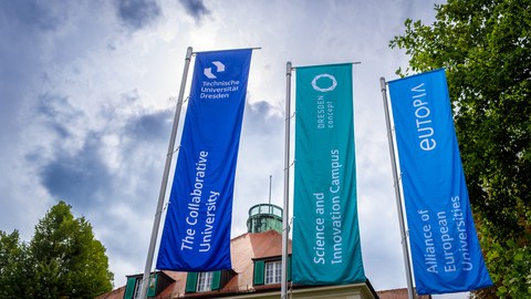 Three flags in front of TU Dresden rectorate with texts 'The Collaborative University', 'Science and Innovation Campus', 'Alliance of European Universities'.