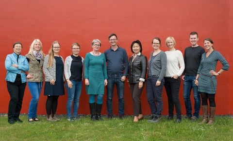 You can see a photo of a group of people. There are eleven people standing on a meadow in front of a red wall. Among them are two men and nine women. It is the team of the Central Student Information and Counselling Service.