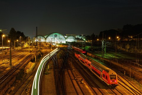 Das Foto zeigt viele Bahnschienen. Auf einer erkennt man einen roten Zug, auf einer anderen lediglich helle Lichtstreifen. Im Hintergrund befindet sich der hell erleuchtete Dresdner Hauptbahnhof.