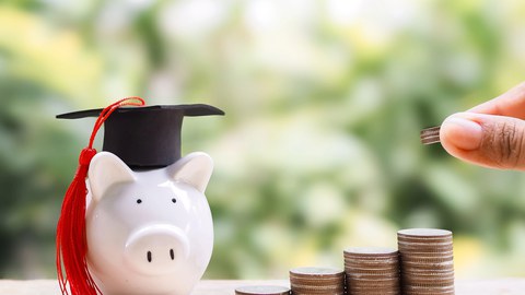 Photo of a piggy bank wearing a doctoral cap. Next to the piggy bank are several large piles of coins, and a hand is holding a few coins.