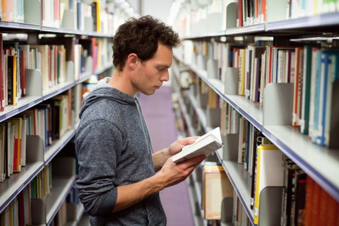 Foto eines Studenten, der zwischen den Bibliotheksregalen steht und ein Buch liest.