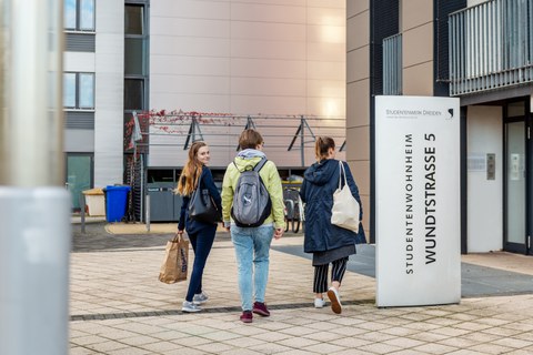 The photo shows three students heading towards the entrance of a student residence. They are carrying rucksacks and bags.