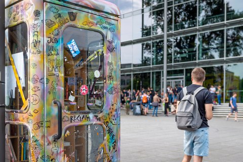 The photo shows a painted telephone box. It is located in front of the lecture hall centre at TU Dresden. There are many books inside the telephone box. Many students are sitting on the steps in front of the lecture hall centre.