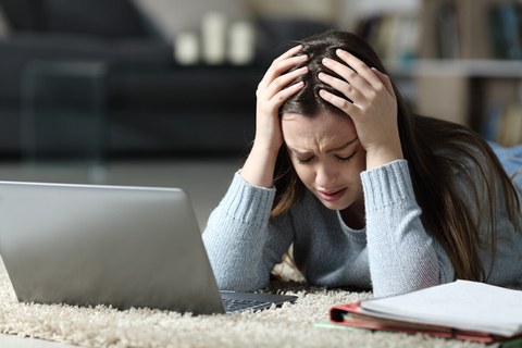 Eine junge Frau liegt verzweifelt vor ihrem Laptop auf dem Teppich, hält ihren Kopf mit beiden Händen und wirkt gestresst. Neben ihr liegt ein aufgeschlagenes Notizbuch mit Unterlagen.