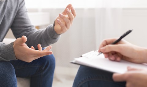 Ein Mann gestikuliert mit den Händen, während er spricht. Gegenüber sitzt eine Person mit Klemmbrett und Stift, die aufmerksam zuhört und Notizen macht – typische Beratungssituation.