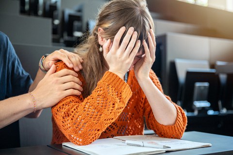 Eine junge Frau sitzt mit den Händen vor dem Gesicht über ihren Unterlagen und wirkt verzweifelt. Neben ihr legt jemand tröstend die Hand auf ihre Schulter, um sie zu unterstützen.