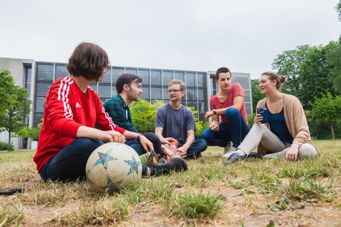 Eine Gruppe junger Studenten sitzt auf einer Wiese und unterhält sich.