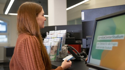 Eine Frau mit roten langen Haaren steht vor einem Schalter des Servicecenter Studium und hält die CampusCard in der Hand.