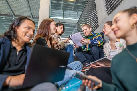 Eine Gruppe Studierender sitzt zusammen auf Treppen, lacht und diskutiert über Notizen und Laptops. Alle wirken konzentriert und arbeiten gemeinsam an einer Aufgabe oder einem Projekt.
