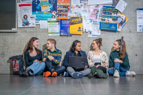 Five young people sit laughing on the floor in front of a bulletin board covered with notices, chatting together.
