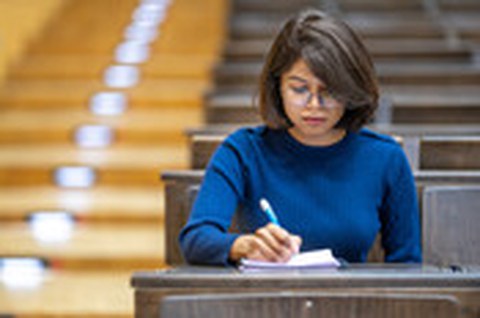 A young woman sits alone in a lecture hall, writing attentively in a notebook.