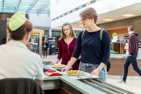 Zwei frauen stehen mit Tabletts und Darauf Tellern mit Essen an einer Kasse. Links im Bild den Rücken der Kassiererin.