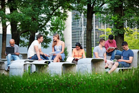 Das Foto zeigt mehrere Studierende draußen im Grünen. Sie sitzen auf einer geschwungenen Betonformation aus Tischen und Bänken und lernen.