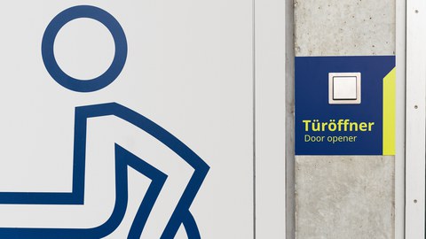Rollstuhlsymbol auf einer Tür. Rechts davon der Schalter eines Türöffners.