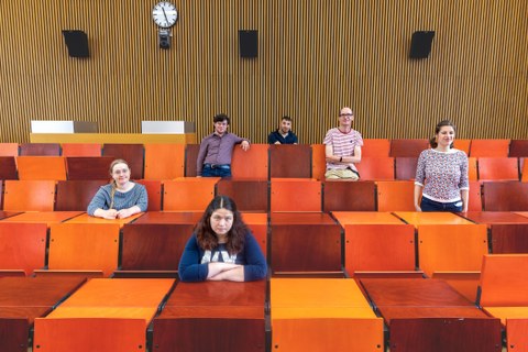 Das Foto zweigt eine Gruppe von 6 Personen, die in einem Vorlesungsaal mit orangenen Sitzgelegenheiten verteilt auf unterschiedliche Reihen sitzen oder stehen. 