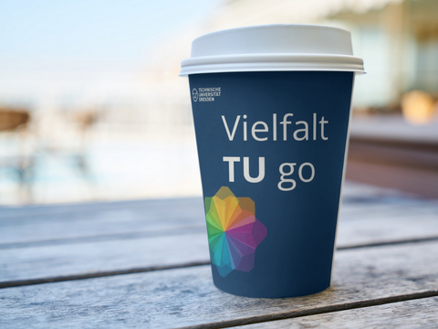 Ein Kaffeebecher mit der Aufschirft "Vielfalt TU go" der auf einer Holzoberfläche steht