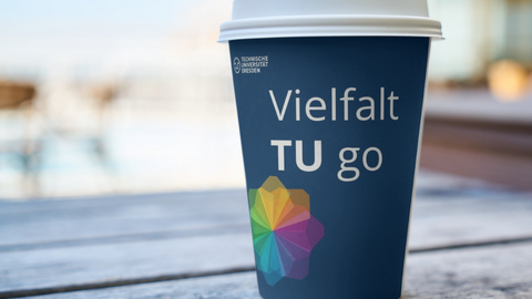 Ein Kaffeebecher mit der Aufschirft "Vielfalt TU go" der auf einer Holzoberfläche steht