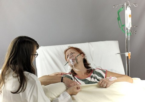 Das Bild zeigt zwei Frauen. Eine Frau liegt in einem Krankenbett und trägt eine Sauerstoffmaske, die andere Frau sitzt neben ihr und hält ihre Hand.