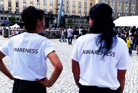 Das Foto zeigt die Rückansicht von 2 Personen. Beide tragen weiße T-Shirts mit dem Rückenaufdruck Awareness in schwarzen Buchstaben.