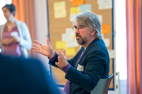 Mann mit grauen Haaren, Brille und blauem Sakko diskutiert