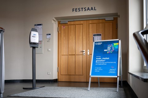 Blaues Infomationsschild vor brauner Tür, welches auf die Veranstaltung hinweist