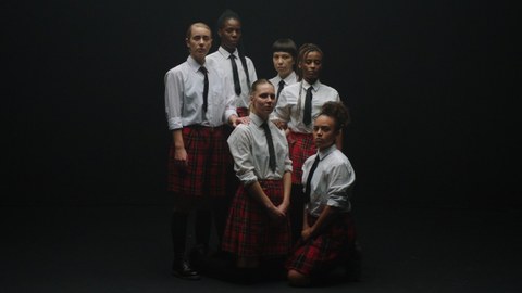 Six young women with different skin colors stand in front of a black background; only the group of people in the middle is lit. They are all wearing the same school uniform, consisting of a white blouse, a dark tie and a knee-high red skirt with a tartan pattern, their calves are covered by dark knee socks. You can see the shoes on some of them, two girls in the foreground have knelt down on the floor so that everyone's faces are clearly visible.