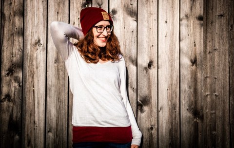 Frau mit weißem Shirt, roten lockigen Haaren und roter Wollmütze vor Holzwand