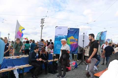 The photo shows people eating and talking together. Beach flags from TU Dresden can be seen next to the tables. 