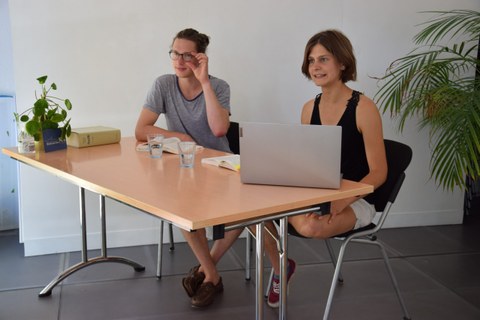 Das foto zeigt zwei junge Personen die an einem Schreibtisch sitzen. Die eine Person hat einen Laptop vor sich aufgeklappt.
