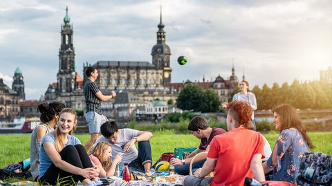 Gruppe von Studierenden an der Elbe. Sie spielen Volleyball und sitzen plaudernd auf der Wiese.