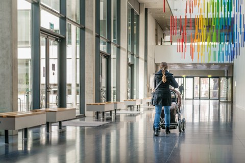 Foto: Eine Frau schiebt einen Kinderwagen über einen Flur mit großen Fenstern im Chemiebau.