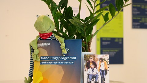 The picture shows a tidy desk or a shelf with several objects on it. In the centre is a catalogue or brochure entitled "Action Programme - Family-friendly University", published by the Technical University of Dresden. To the left of the brochure sits a gr