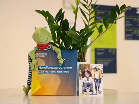 The picture shows a tidy desk or a shelf with several objects on it. In the centre is a catalogue or brochure entitled "Action Programme - Family-friendly University", published by the Technical University of Dresden. To the left of the brochure sits a gr