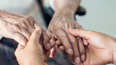 Das Foto zeigt die faltigen Hände einer älteren Person, welche in den Händen einer jüngeren Person liegen und von diesen umschlossen werden.