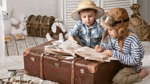 Das Foto zeigt zwei Kinder. Sie haben Forscherhüte auf und sitzen vor einem großen braunem Koffer. Auf dem Koffer liegt ein aufgeschlagenes Reisetagebuch. Ein Kind hält eine Feder in der Hand, mit der er gerade etwas in das Buch schreibt.