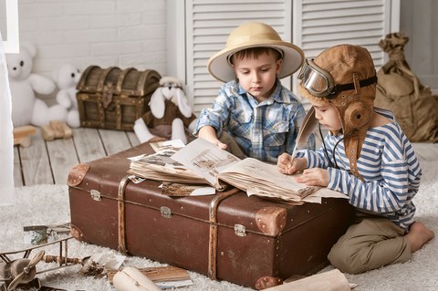 Das Foto zeigt zwei Kinder. Sie haben Forscherhüte auf und sitzen vor einem großen braunem Koffer. Auf dem Koffer liegt ein aufgeschlagenes Reisetagebuch. Ein Kind hält eine Feder in der Hand, mit der er gerade etwas in das Buch schreibt.