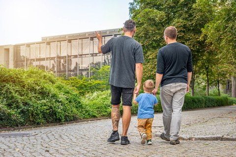 Zwei männlich gelesene Personen gehen mit einem Kind durch eine grüne, städtische Umgebung.